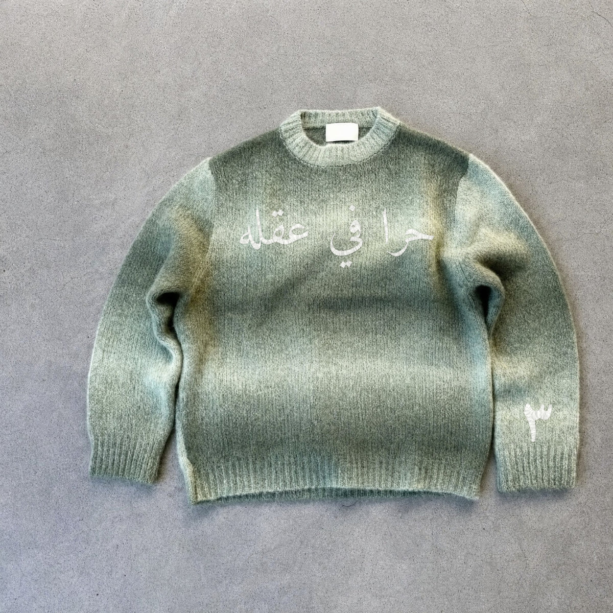 حرا Strickpullover in Salbeigrün, Vorderseite mit weißer Stickerei «حرا في عقله»
