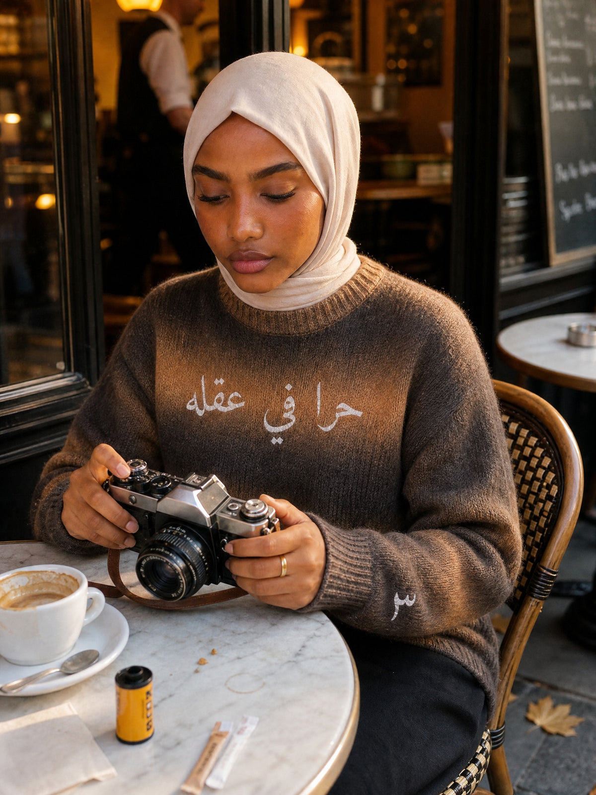 Amina im SYRVAIN «حرا» Strickpullover Braun mit Filmkamera im Café in Paris